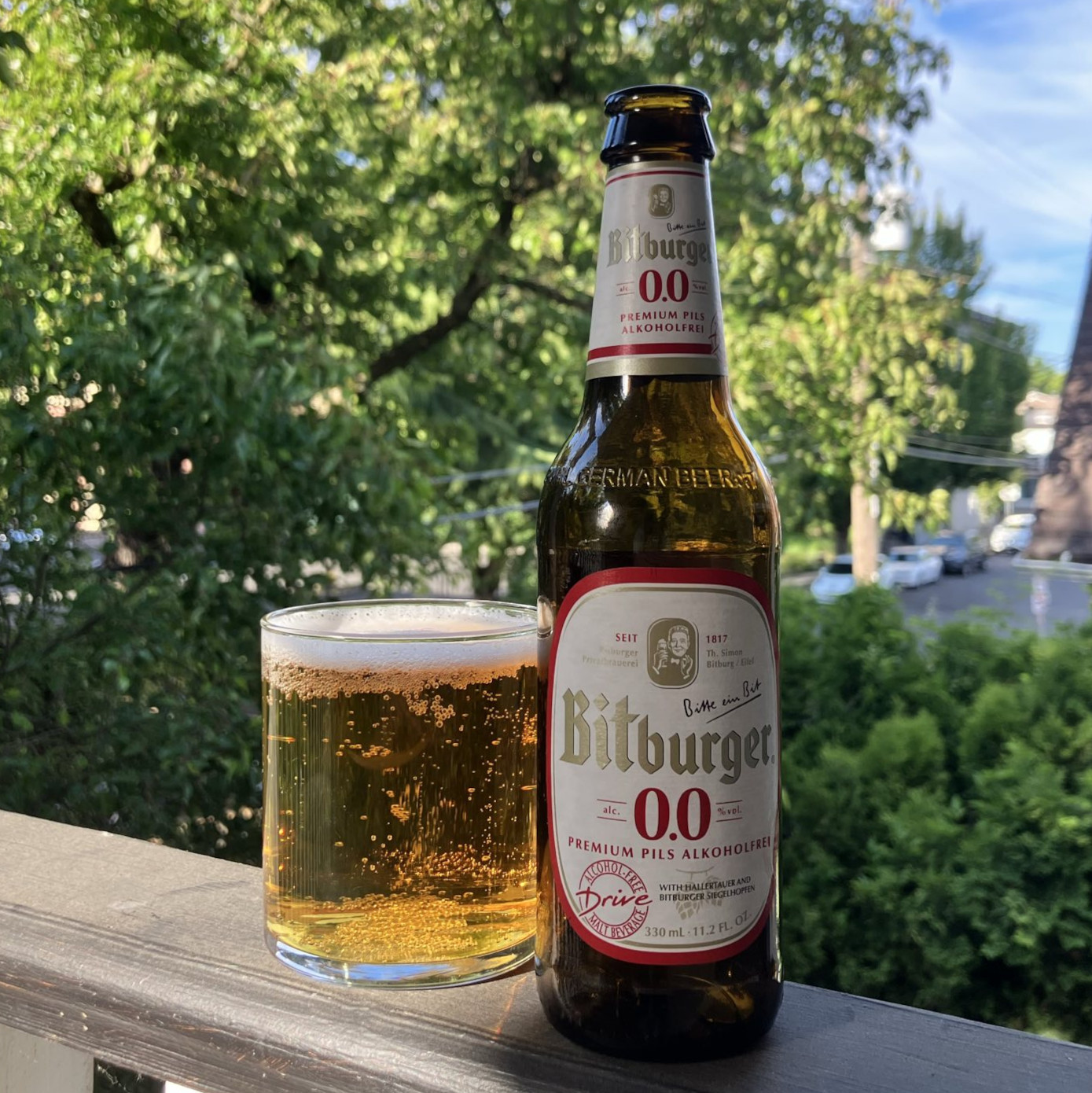 Bitburger&rsquo;s Bitburger 0.0 in a 330 milliliter bottle, poured into a minimalist lowball glass, on a balcony railing